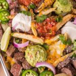 Tray of loaded carne asada fries topped with steak, guacamole, salsa, sour cream, jalapeños, red onion, shredded cheese, cilantro, and lime wedges.