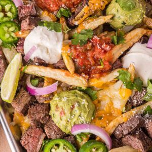 Tray of loaded carne asada fries topped with steak, guacamole, salsa, sour cream, jalapeรฑos, red onion, shredded cheese, cilantro, and lime wedges.
