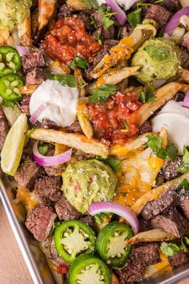 Tray of loaded carne asada fries topped with steak, guacamole, salsa, sour cream, jalapeños, red onion, shredded cheese, cilantro, and lime wedges.