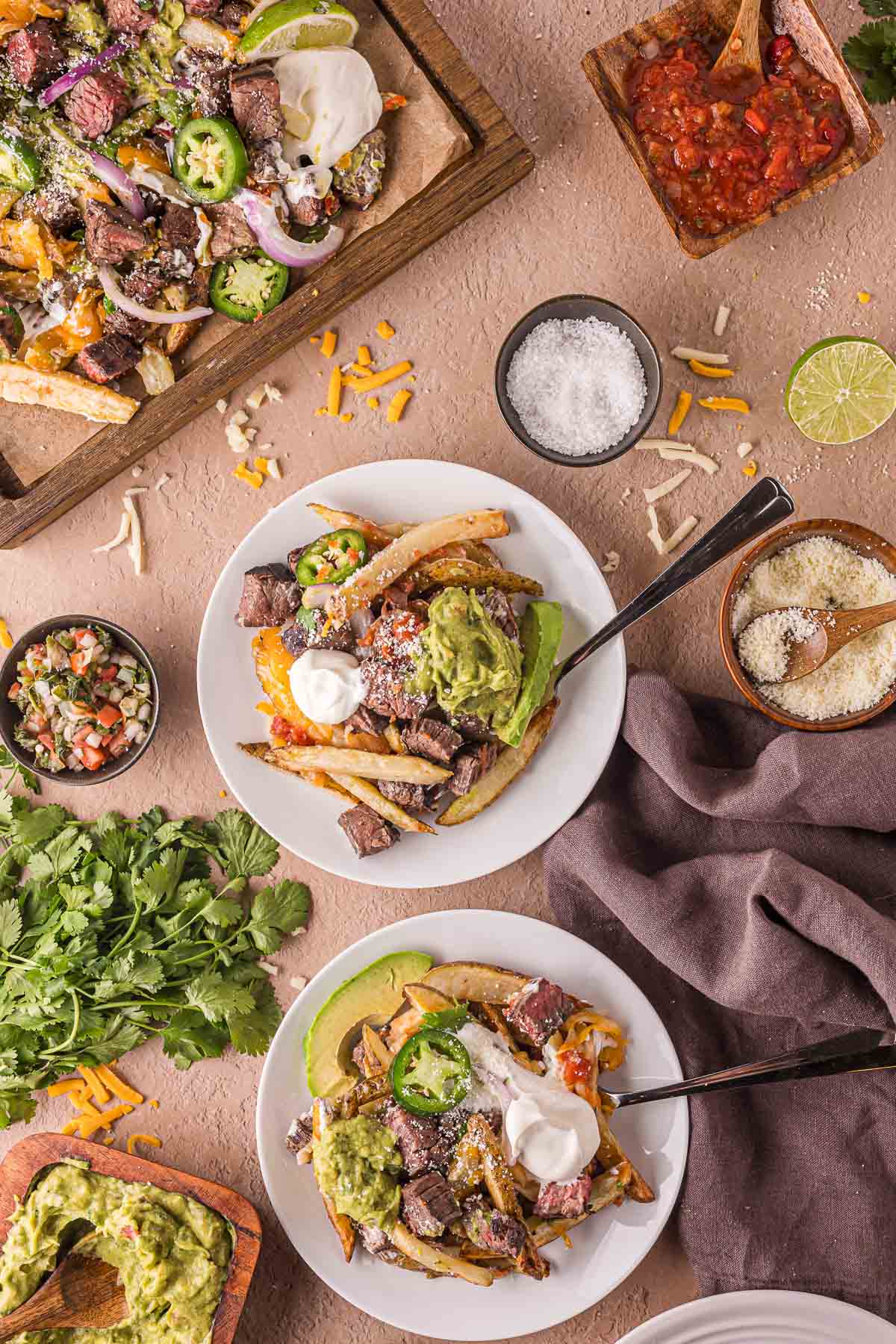 Two plates of carne asada fries topped with guacamole, sour cream, and cheese, surrounded by bowls of salsa, grated cheese, lime, cilantro, and other toppings on a beige surface.