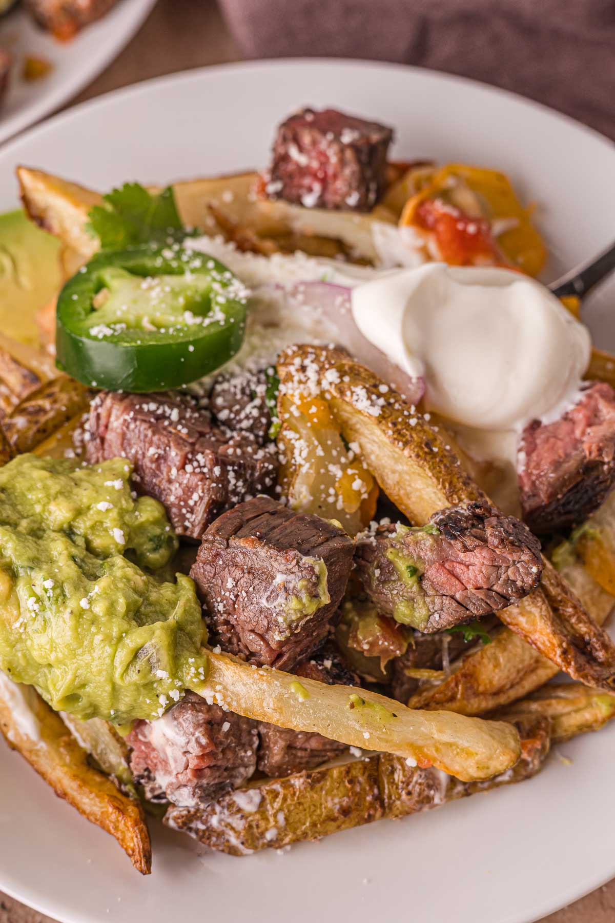 A plate of carne asada steak fries topped with grilled steak pieces, guacamole, sour cream, sliced jalapeรฑo, shredded cheese, and pico de gallo.