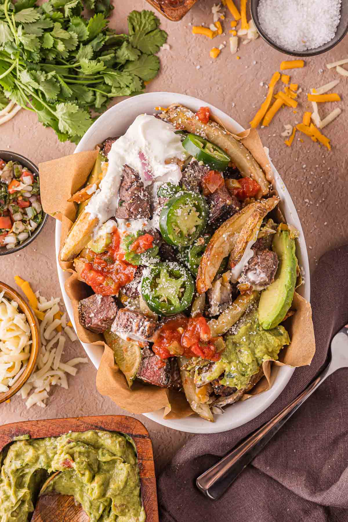 A bowl filled with carne asada steak fries topped with sour cream, guacamole, avocado slices, jalapeรฑos, shredded cheese, salsa, and chopped vegetables, on a brown-paper lining.