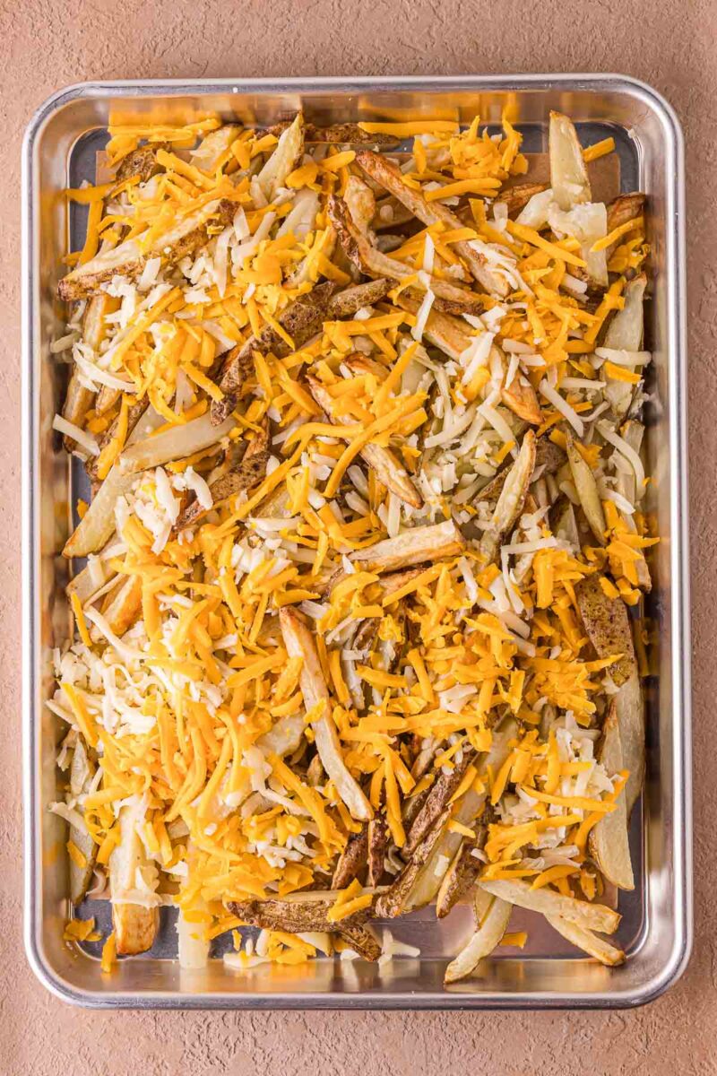 A baking tray filled with cooked French fries topped with shredded yellow and white cheese.