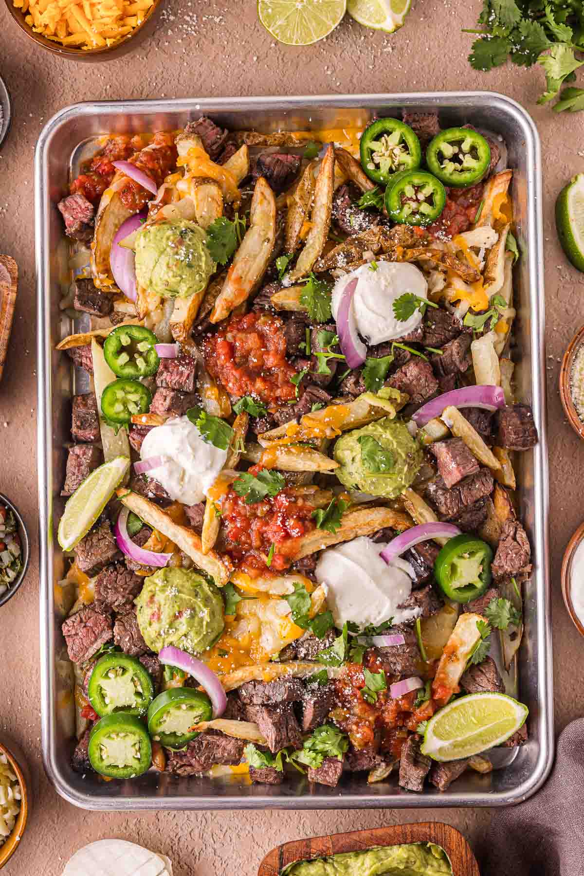 A metal tray filled with loaded carne asada fries topped with cheese, guacamole, sour cream, salsa, jalapeรฑos, red onion, cilantro, and lime wedges.