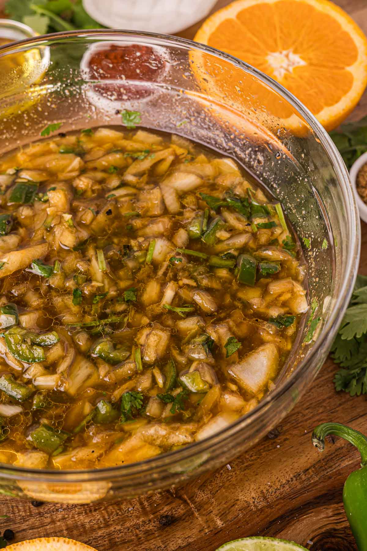 A glass bowl filled with a chunky carne asada marinade containing chopped onions, green peppers, and herbs, surrounded by orange slices and small bowls of spices.