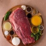 Raw flank steak on a wooden board, surrounded by small bowls of spices, sauces, broth, chopped herbs, a chili pepper, garlic bulb, and a white onion.
