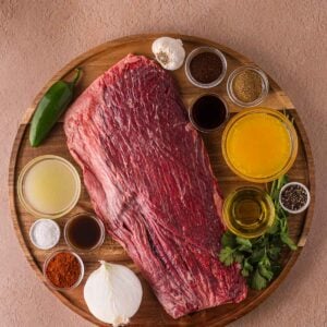 Raw flank steak on a wooden board, surrounded by small bowls of spices, sauces, broth, chopped herbs, a chili pepper, garlic bulb, and a white onion.