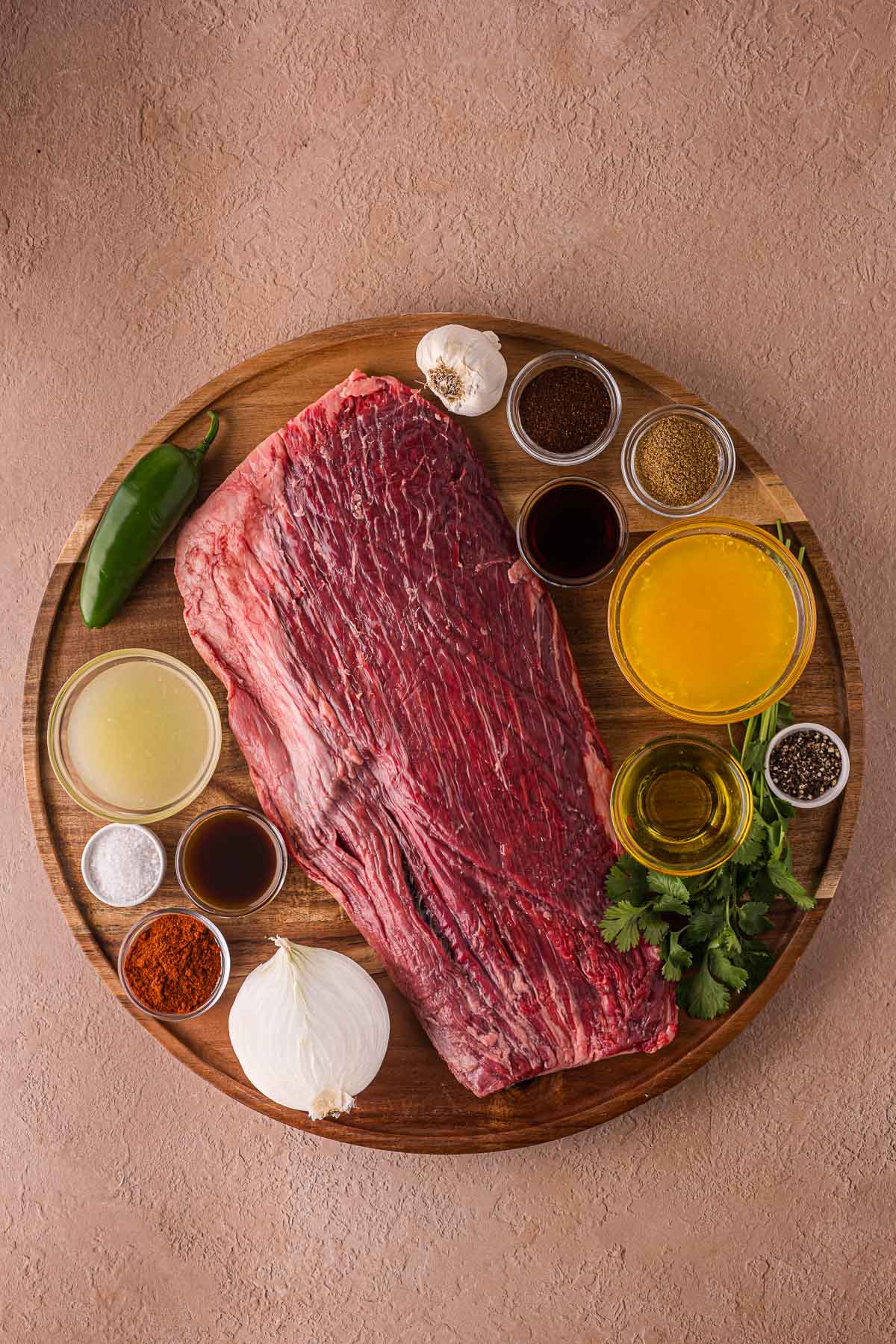 Raw flank steak on a wooden board, surrounded by small bowls of spices, sauces, broth, chopped herbs, a chili pepper, garlic bulb, and a white onion.