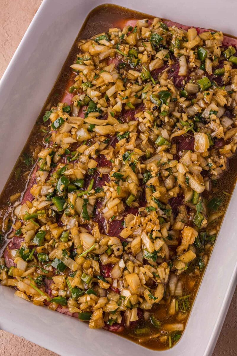 A white dish containing raw meat covered with a carne asada marinade made of chopped onions, herbs, and green peppers.