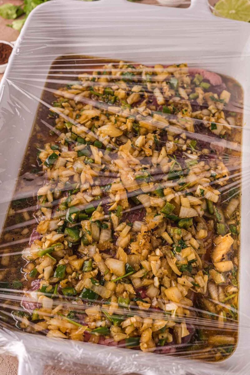 A white dish covered with plastic wrap contains meat marinating in carne asada marinade.