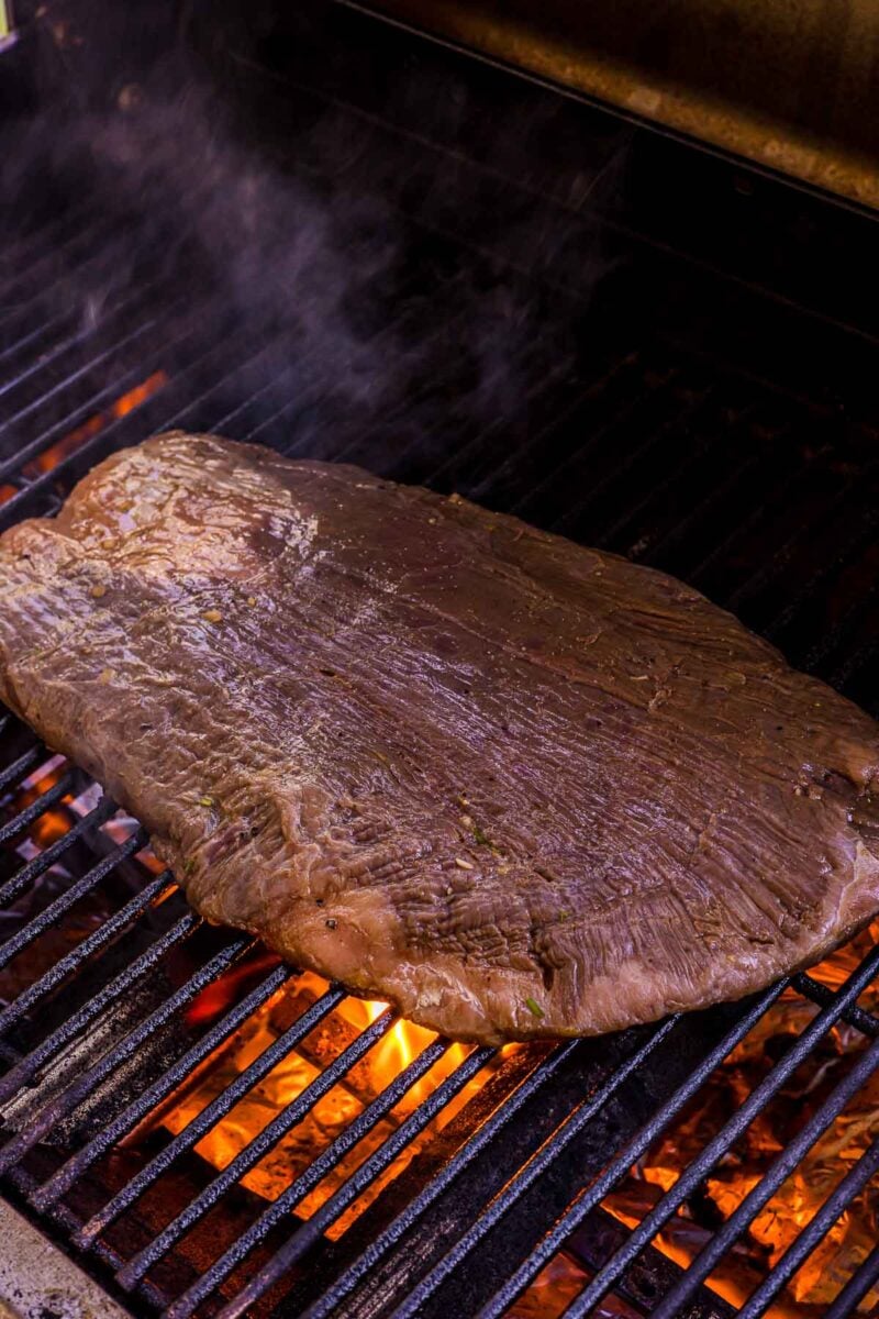 A large piece of marinated flank steak is being grilled over open flames on a barbecue grill.