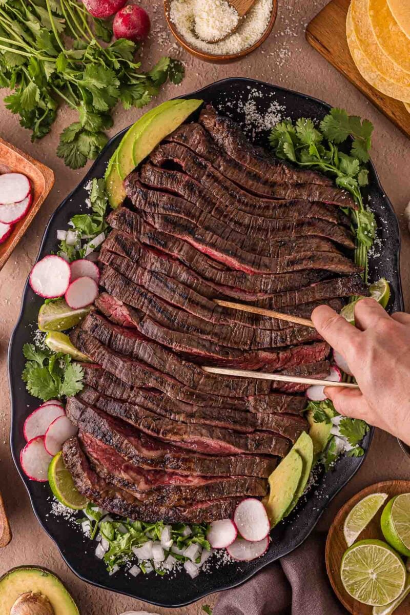 Sliced grilled carne asada arranged on a black platter with avocado, radish, cilantro, and lime; a hand is cutting the meat.