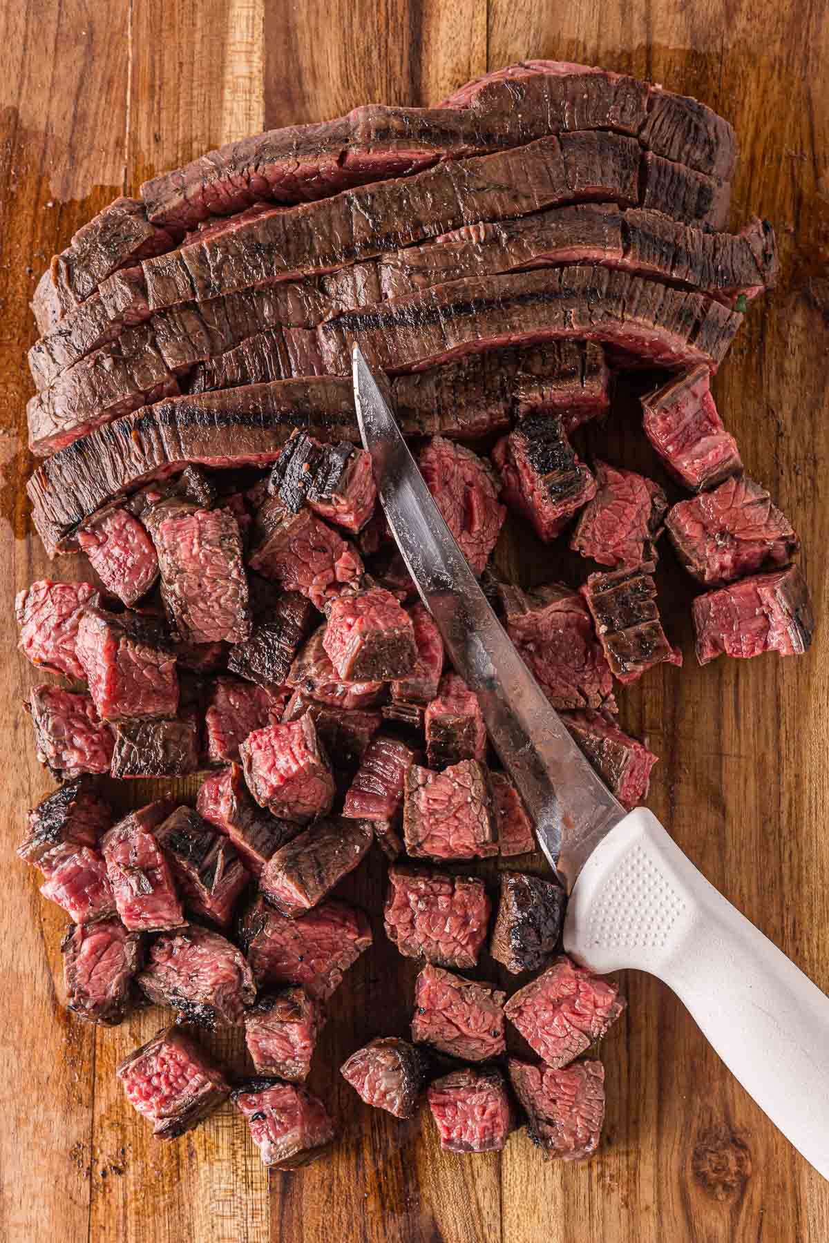 Sliced carne asada being cut into small bites on a wooden cutting board with a knife.