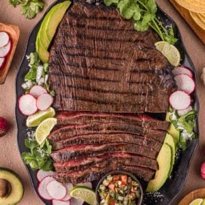 A large platter of grilled carne asada served with sliced avocado, radishes, lime wedges, cilantro, and a small bowl of salsa, surrounded by tortillas and additional garnishes.