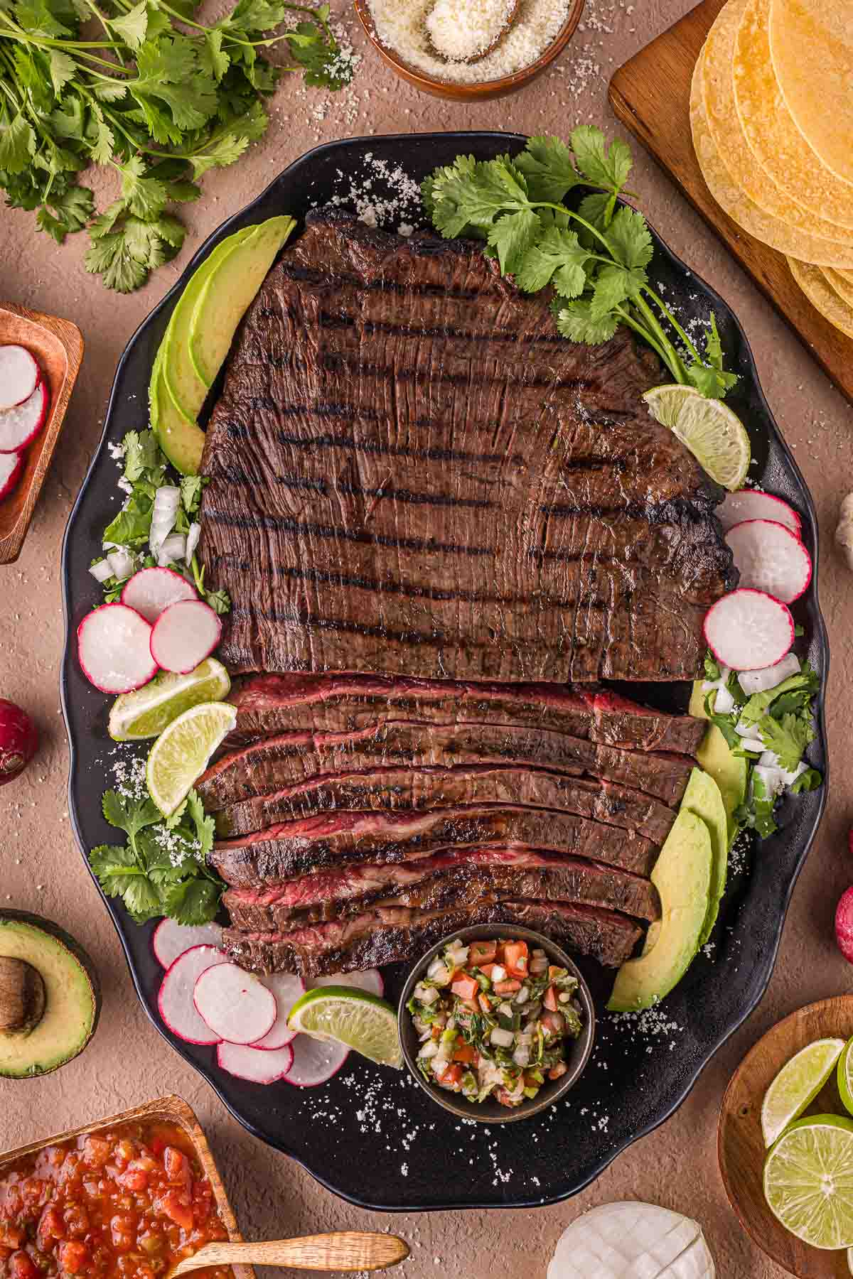 A large platter of grilled carne asada served with sliced avocado, radishes, lime wedges, cilantro, and a small bowl of salsa, surrounded by tortillas and additional garnishes.