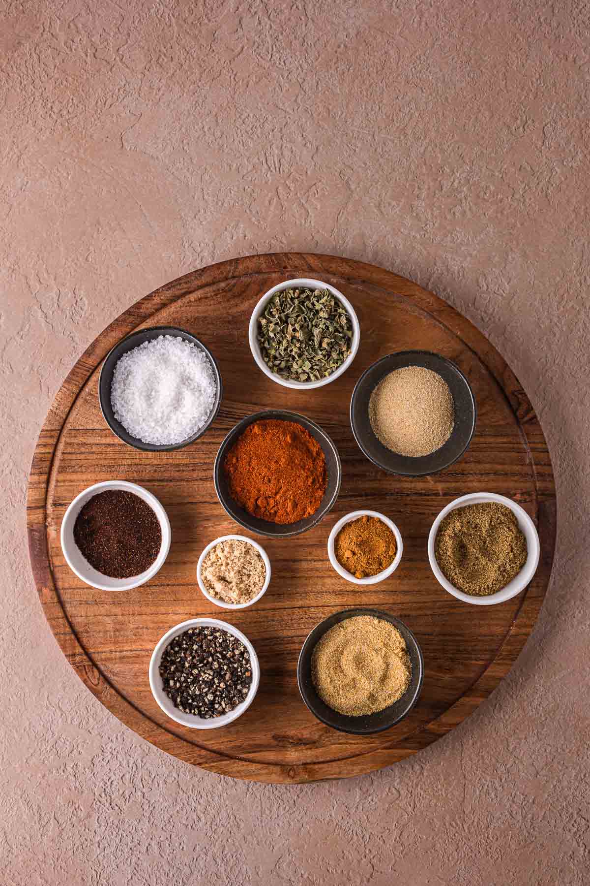 A round wooden board holds ten small bowls filled with assorted spices and seasonings for a homemade carne asada rub, arranged in a circular pattern on a textured surface.