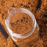 A glass jar filled with a carne asada spice mix is surrounded by more of the same spice spread on a dark surface.