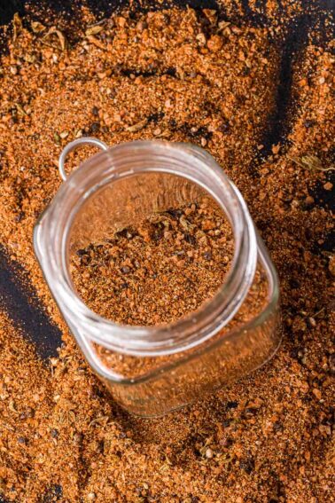 A glass jar filled with a carne asada spice mix is surrounded by more of the same spice spread on a dark surface.