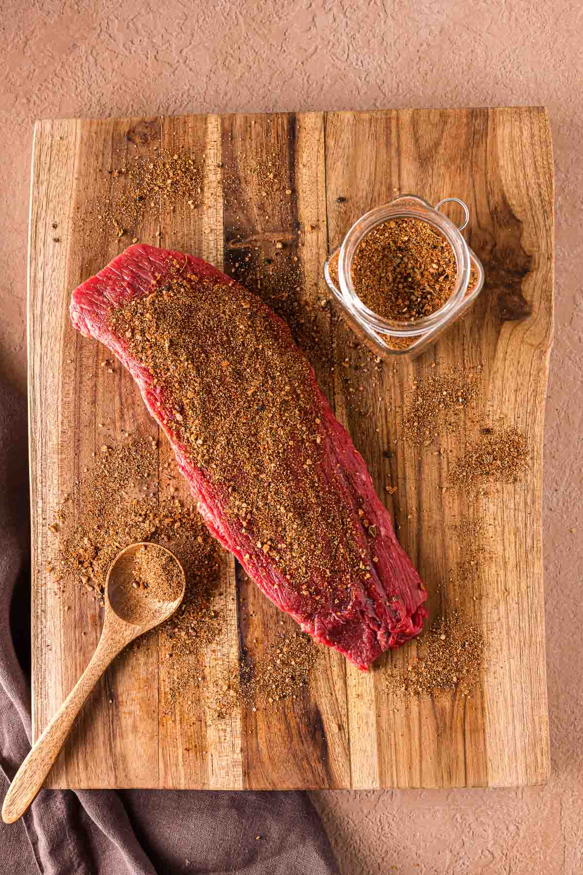A raw cut of beef sprinkled with dry carne asada spice rub sits on a wooden cutting board next to a wooden spoon and a small jar of seasoning.