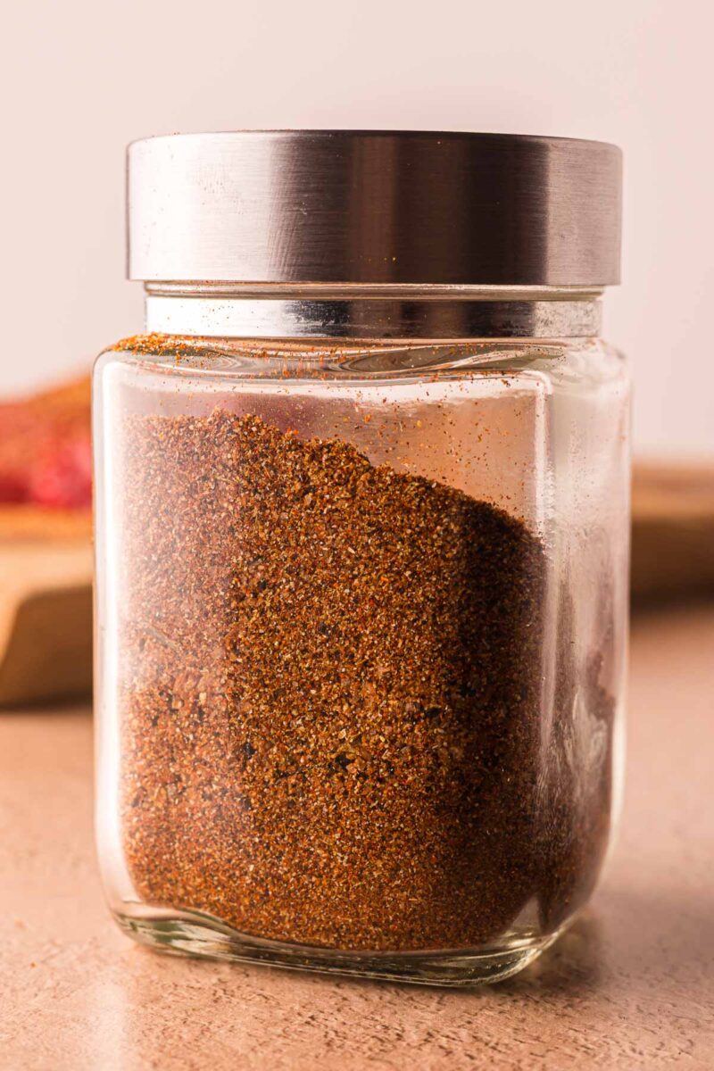 A clear glass jar with a metal lid filled with a reddish-brown carne asada seasoning blend, sitting on a light countertop.