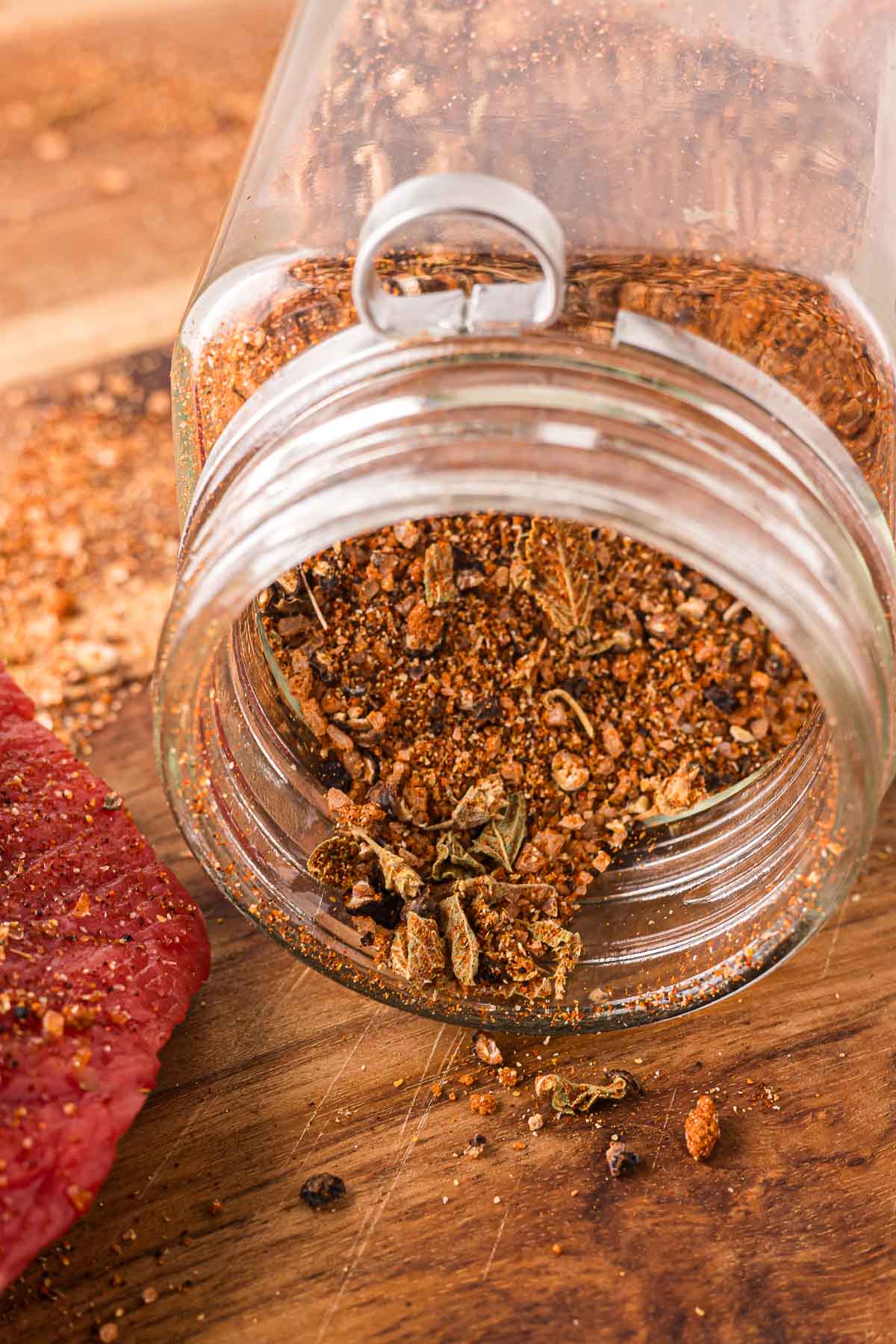 A glass jar of carne asada seasoning spilled on a wooden surface next to a piece of raw red meat.