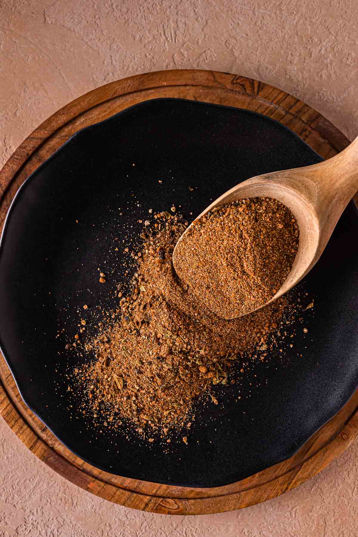 A wooden spoon holds and sprinkles a brown carne asada seasoning spice blend onto a black plate set on a wooden charger against a light textured surface.