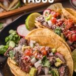Two carne asada tacos topped with diced onions, cilantro, and salsa are served on a black plate with lime wedges and radish slices.