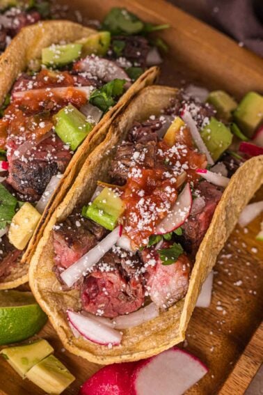 Two carne asada tacos in corn tortillas topped with diced avocado, radish slices, onions, salsa, cilantro, and crumbled cheese, served on a wooden board.