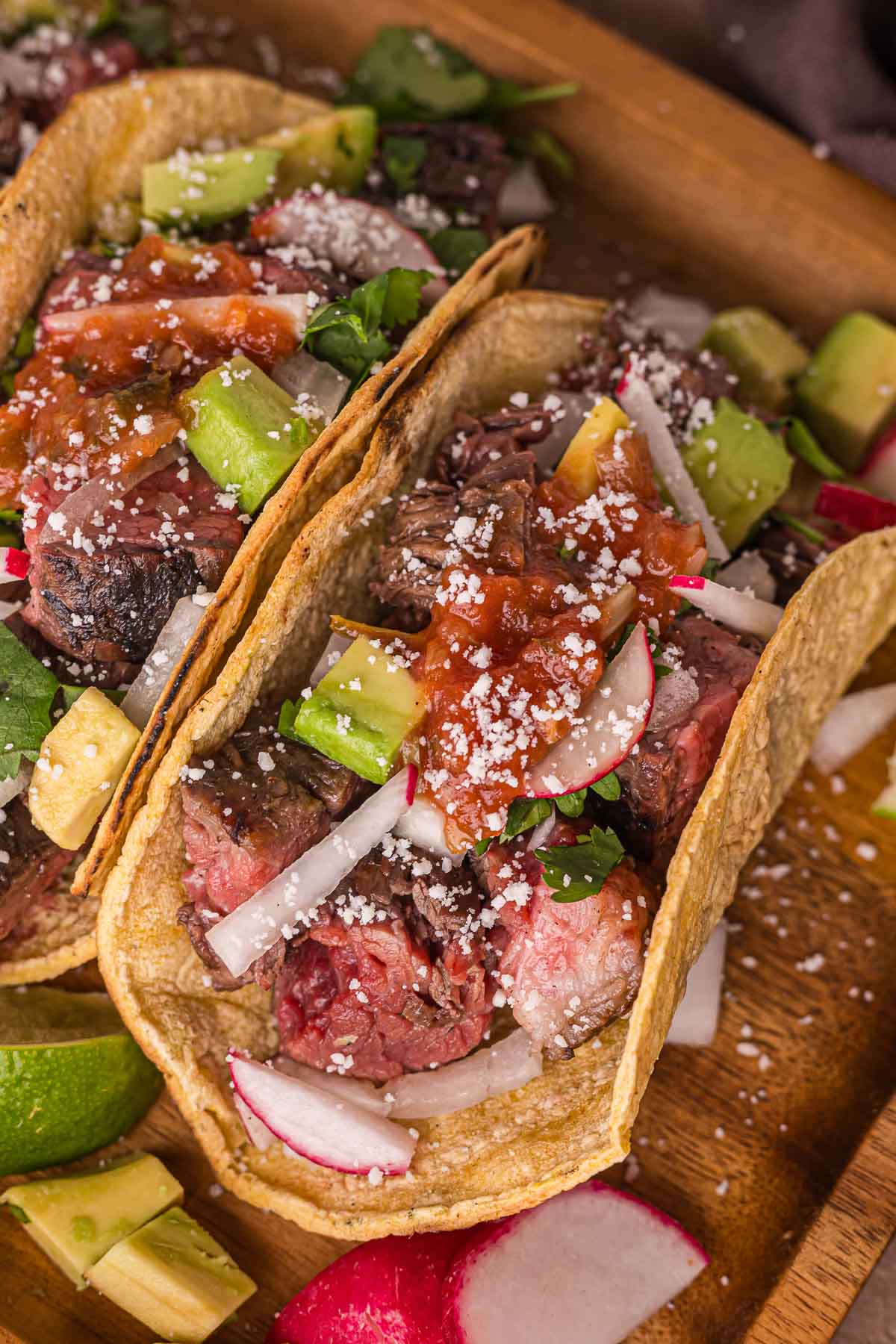 Two carne asada tacos in corn tortillas topped with diced avocado, radish slices, onions, salsa, cilantro, and crumbled cheese, served on a wooden board.