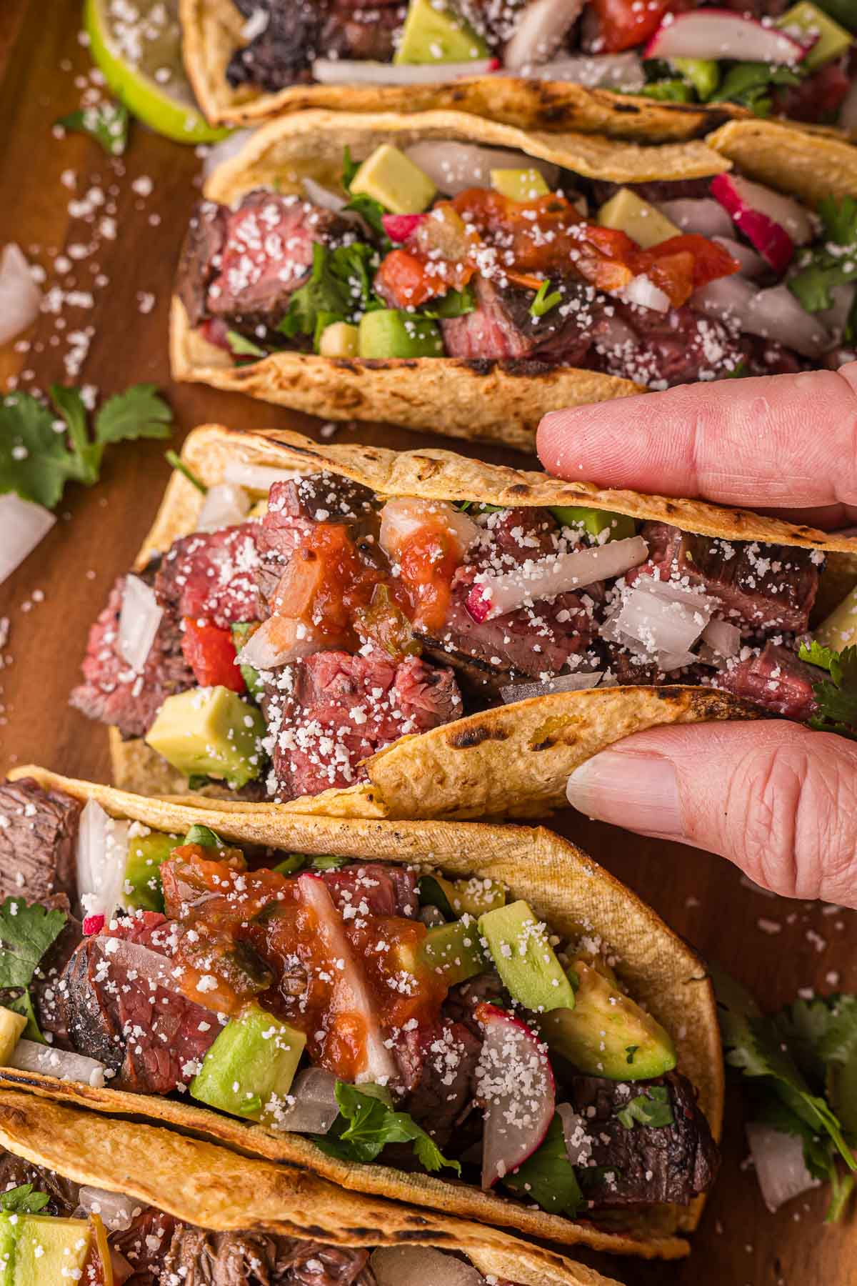 A hand holding a carne asada taco topped with diced onions, avocado, tomato salsa, radishes, and crumbled cheese, alongside similar tacos on a wooden surface.