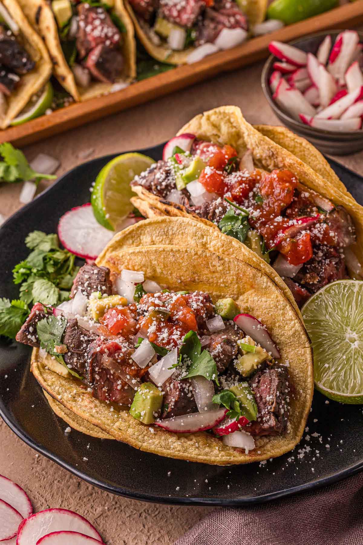 Two carne asada steak tacos on corn tortillas topped with diced onions, cilantro, salsa, avocado, radish slices, and crumbled cheese, served with lime wedges on a black plate.