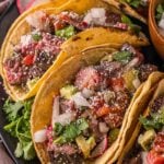Steak tacos filled with diced steak, onions, cilantro, avocado, salsa, and cheese on corn tortillas, garnished with lime wedges and fresh cilantro on a dark plate.