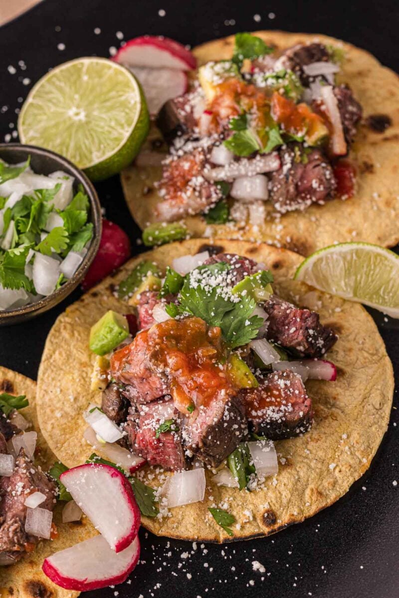 Carne asada steak tacos topped with chopped onions, cilantro, salsa, and cheese are served on corn tortillas with lime wedges and sliced radishes on a black plate.