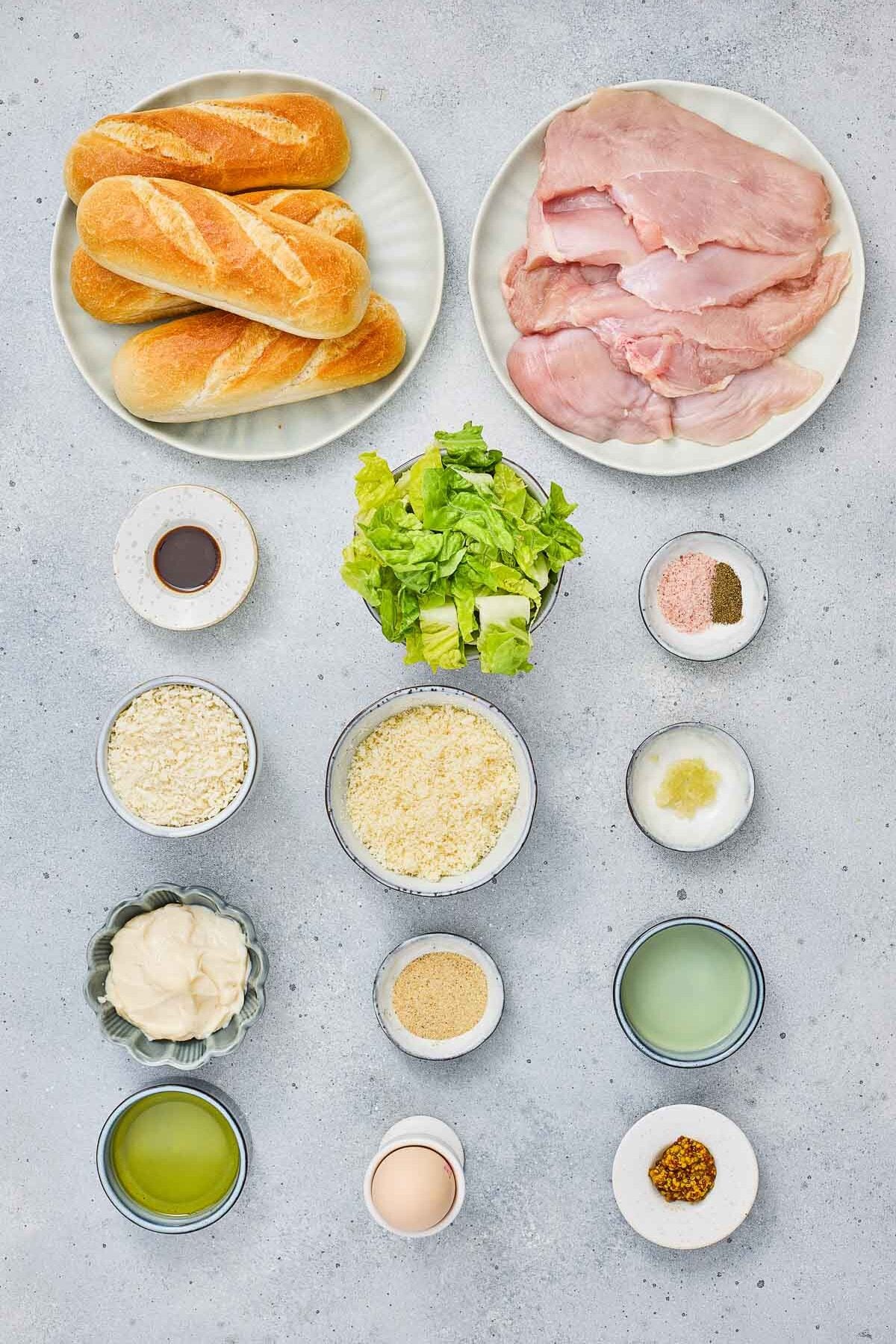 Overhead view of ingredients for crispy caesar chicken sandwiches on a gray surface, including rolls, raw chicken breast, lettuce, seasonings, sauces, egg, oil, and breadcrumbs arranged in bowls and plates.