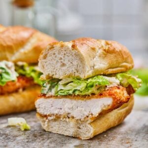 A crispy caesar chicken sandwich with lettuce and sauce, cut in half and displayed on a stone surface with a blurred background.