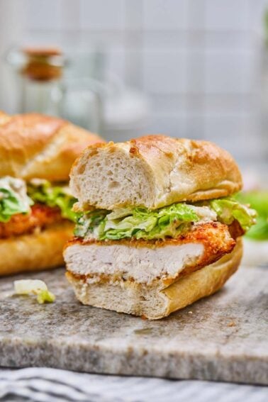 A crispy caesar chicken sandwich with lettuce and sauce, cut in half and displayed on a stone surface with a blurred background.