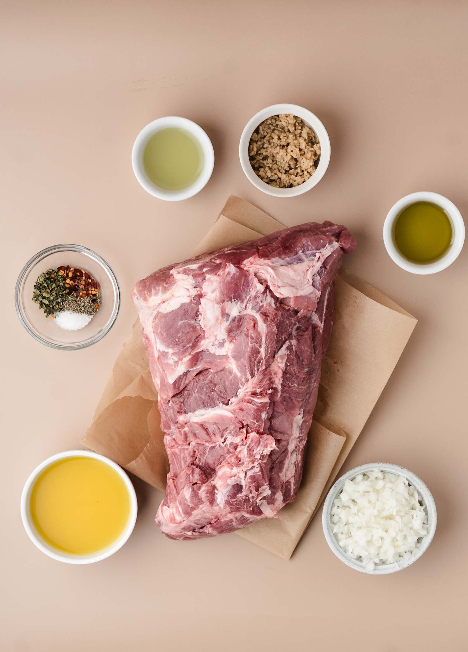 Raw pork shoulder on parchment paper, surrounded by bowls of olive oil, chopped onions, brown sugar, herbs and spices, broth, and another liquid ingredient on a beige surface.