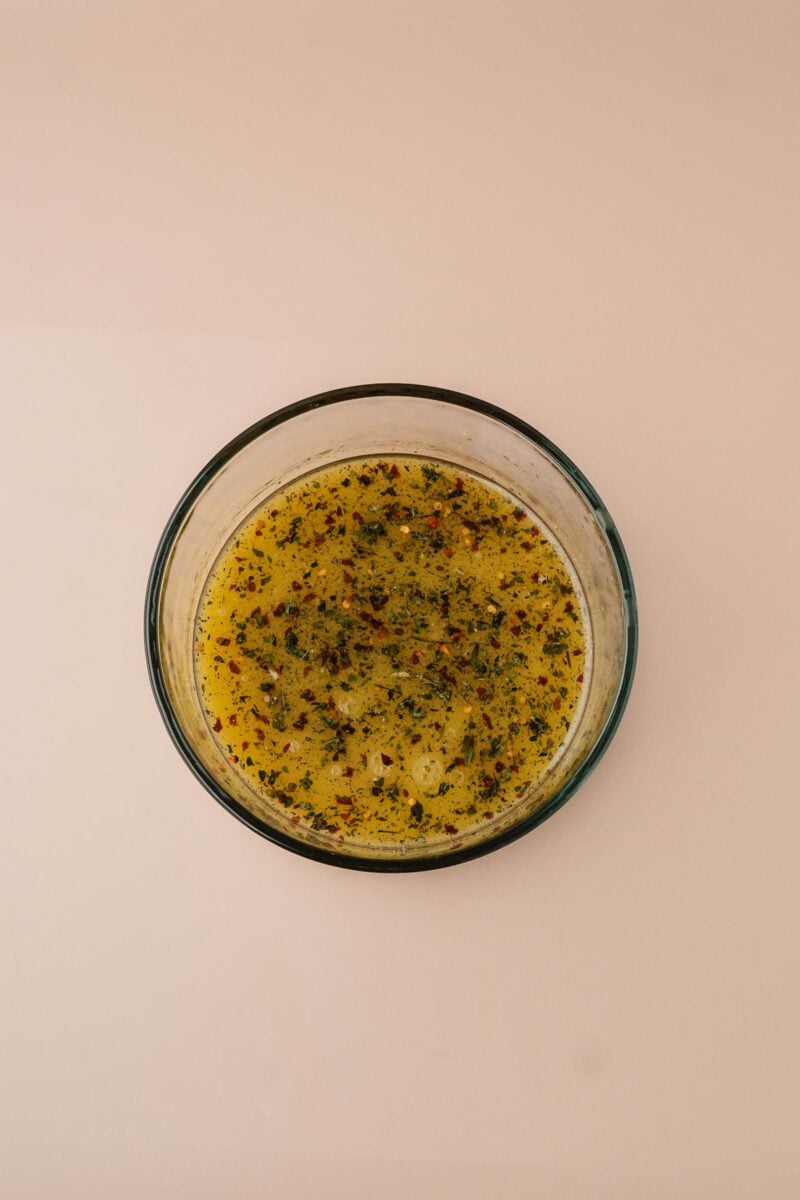 A glass bowl filled with a yellow, oily marinade seasoned with herbs and spices, placed on a plain light-colored surface.