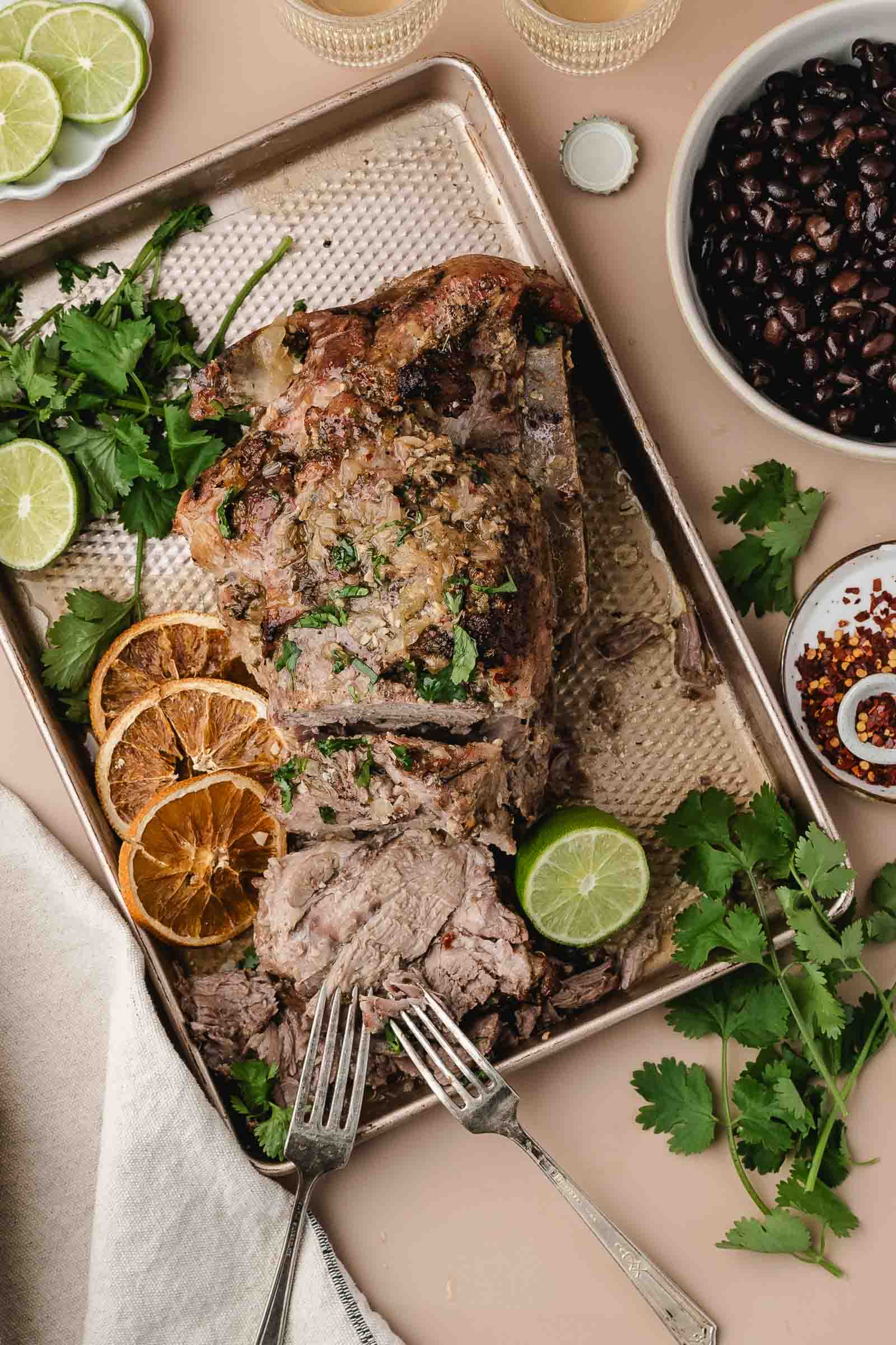 A baked mojo pork roast garnished with cilantro and lime, served on a tray with two forks starting to shred it and sliced dried oranges, fresh cilantro, lime halves, and bowls of black beans and red pepper flakes nearby.