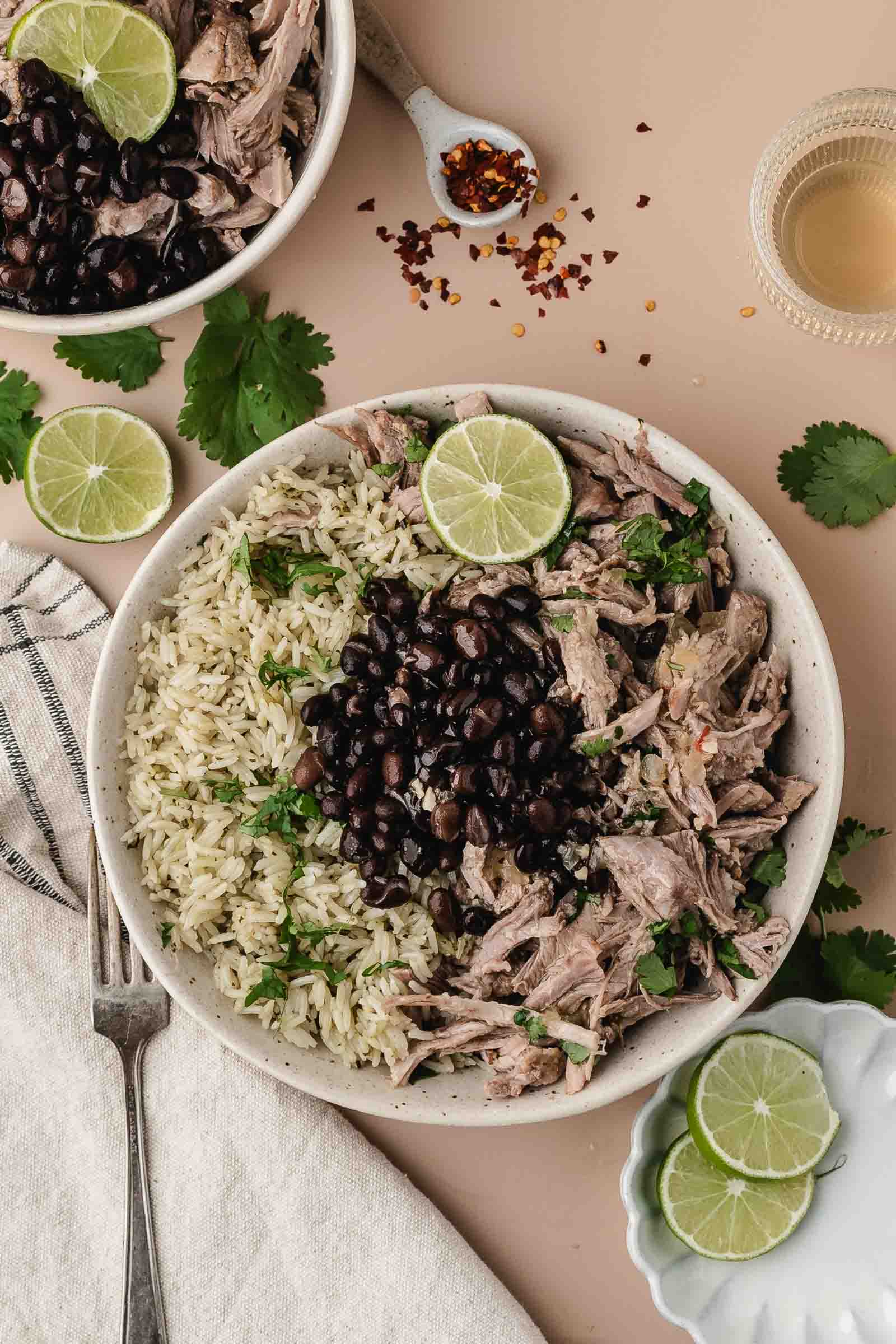 A bowl with shredded mojo pork, black beans, cilantro rice, and a lime slice, surrounded by cilantro leaves, lime wedges, a drink, and a napkin on a beige surface.