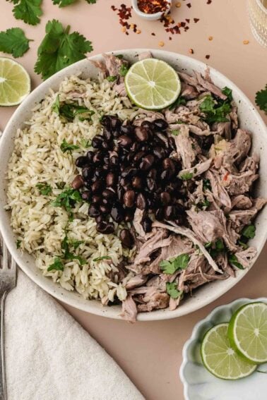 A bowl of cilantro rice, shredded mojo pork, and black beans garnished with fresh cilantro and lime slices.