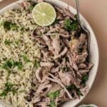 A bowl containing shredded mojo pork garnished with cilantro and lime, served alongside a portion of rice. A spoon rests in the bowl; extra lime slices and beans are nearby.