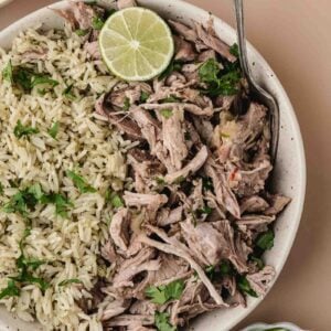 A bowl containing shredded mojo pork garnished with cilantro and lime, served alongside a portion of rice. A spoon rests in the bowl; extra lime slices and beans are nearby.
