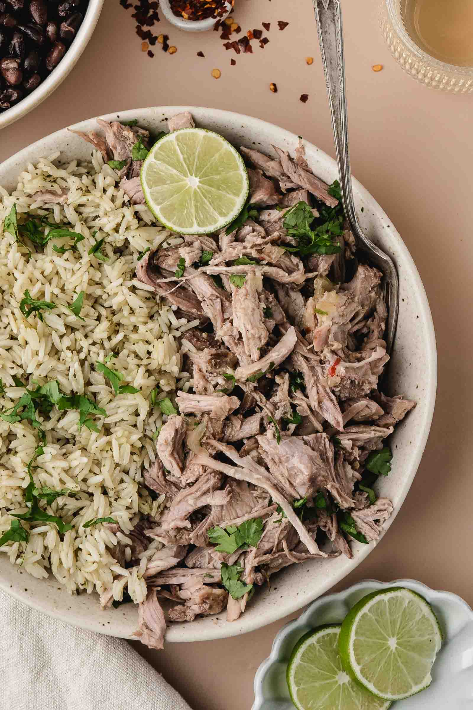 A bowl containing shredded mojo pork garnished with cilantro and lime, served alongside a portion of rice. A spoon rests in the bowl; extra lime slices and beans are nearby.