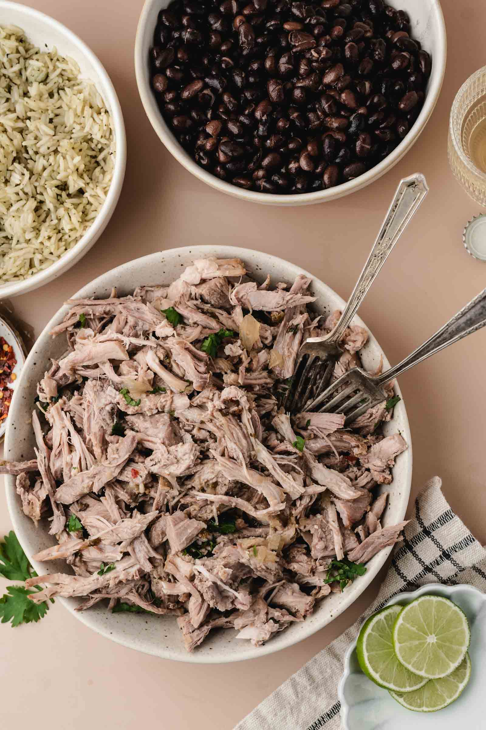 A bowl of shredded Cuban mojo pork with two forks beside bowls of black beans, rice, a drink, lime slices, and a napkin on a light surface.