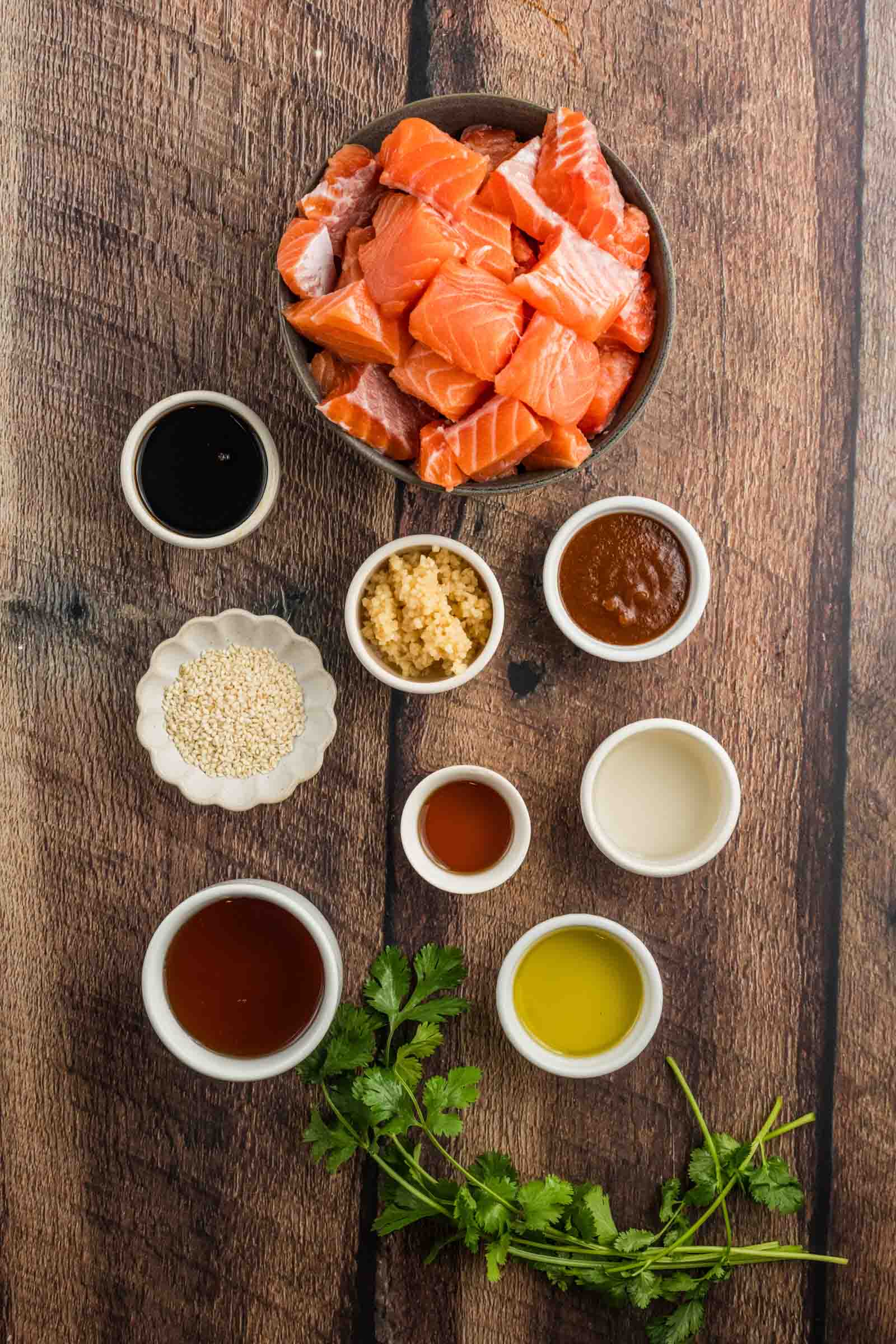 A bowl of raw salmon cubes surrounded by small bowls of sauces, minced garlic, sesame seeds, and fresh cilantro on a wooden surface.