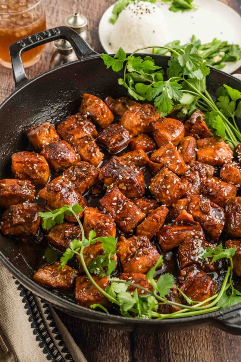A skillet filled with glazed, seared salmon bites garnished with fresh cilantro, with a plate of rice and herbs in the background.