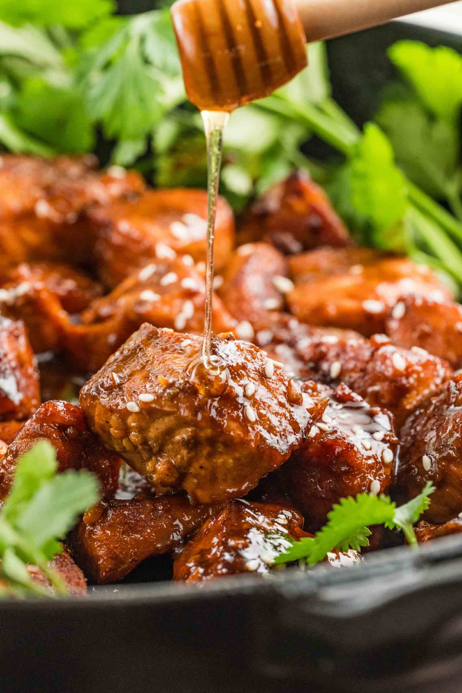Close-up of glazed salmon pieces garnished with sesame seeds and cilantro, with honey being drizzled over them.