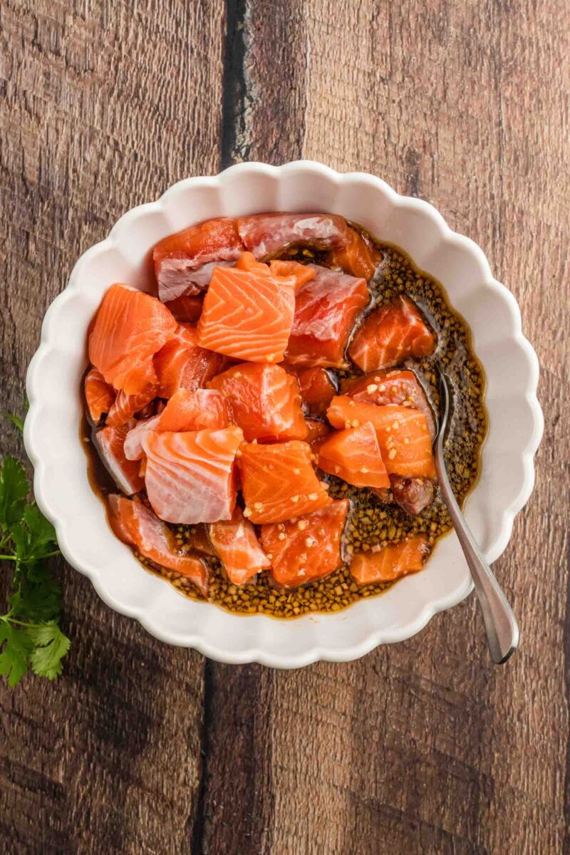 A white bowl with raw salmon chunks marinating in a dark sauce, with a spoon inside, placed on a wooden surface.