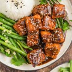 Plate with white rice topped with black sesame seeds, green beans, and glazed salmon bites garnished with sesame seeds, on a wooden table.